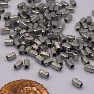 May include: A large quantity of silver-toned metal beads, each with a cylindrical shape and a small hole at each end. The beads are scattered on a white surface, with a US penny coin in the lower right corner for scale.