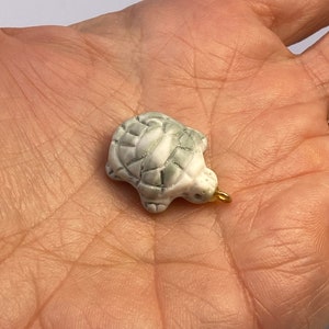 May include: A small white ceramic turtle charm with a green glaze. The turtle has a gold loop for attaching to a necklace or bracelet.