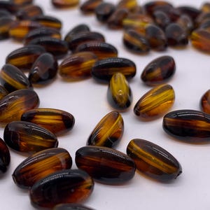 May include: A collection of brown and amber oval-shaped glass beads. The beads have a striped pattern and are scattered on a white surface.