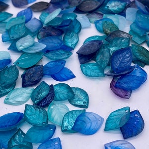 May include: A collection of blue, teal, and purple iridescent glass leaf-shaped charms. The charms have a shiny, glittery finish and are perfect for jewelry making or crafting.