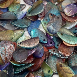 Könnte beinhalten: Eine große Auswahl an schimmernden, glitzernden Perlen in Blattform in verschiedenen Blautönen, Grüntönen, Violetttönen und Brauntönen. Die Perlen haben eine strukturierte, leicht erhabene Oberfläche und sind perfekt für die Schmuckherstellung oder das Basteln.