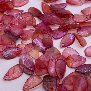 May include: A pile of pink and purple iridescent leaf-shaped beads with a hole at the top for stringing. The beads have a subtle glitter finish.