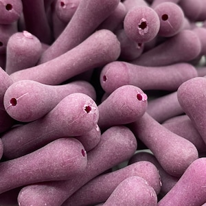 May include: A close-up of a pile of purple, smooth, rounded, ceramic beads. The beads are shaped like small, elongated teardrops with a hole in the center.