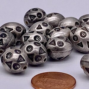 May include: A collection of silver-toned beads with geometric patterns, including circles and triangles. The beads are oval-shaped and have a slightly textured surface.