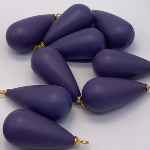 May include: A collection of teardrop-shaped beads in a deep purple hue. Each bead has a matte finish and is topped with a small gold-colored loop. The beads are scattered against a white background, suggesting they are for jewelry making or crafting.