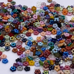 May include: A collection of small, flower-shaped beads in various colors, including blue, pink, green, and gold. The beads are scattered across a white surface, creating a colorful and textured display. These beads are ideal for jewelry making and craft projects.