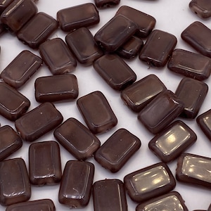 May include: A group of dark brown rectangular glass beads. The beads have a glossy finish and are scattered on a white surface.