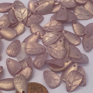 Könnte beinhalten: Eine Sammlung von rosa und goldenen, blattförmigen Perlen mit einer strukturierten Oberfläche. Die Perlen haben oben ein Loch zum Auf fädeln.