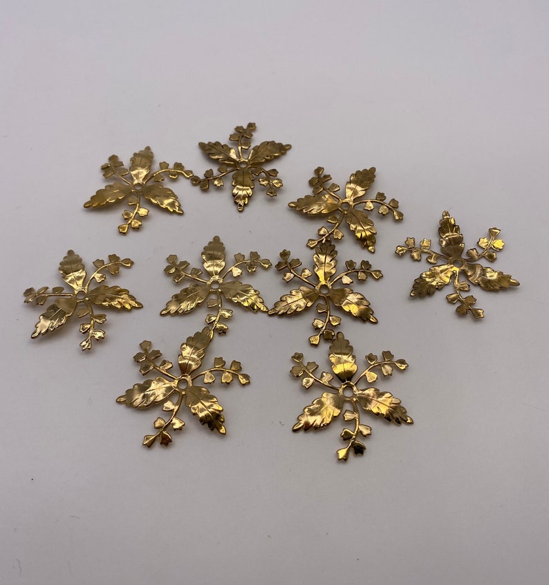 May include: Ten gold metal floral embellishments with a detailed leaf design. Each embellishment has a small hole in the center.