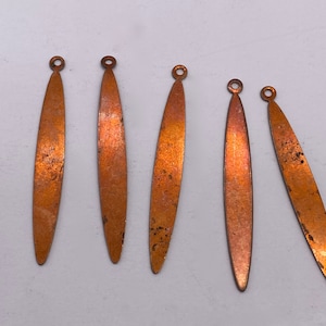 May include: Five copper-colored metal pendants with a loop at the top. The pendants are oval-shaped and have a slightly textured surface.