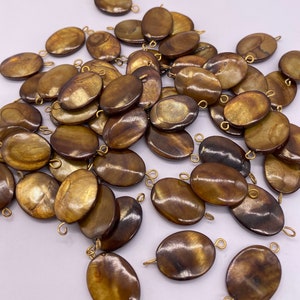 Puede incluir: Un conjunto de colgantes ovalados marrones iridiscentes con bucles dorados para ensartar. Los colgantes tienen un acabado pulido y brillante, y varían en tonos marrones y dorados. Estos colgantes son adecuados para la fabricación de joyas.