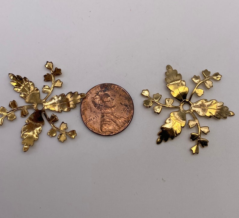 May include: Two gold-colored metal decorative pieces shaped like a stylized fern with a hole in the center. The pieces are about the size of a US penny.