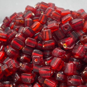 Może przedstawiać: Stosik małych, kwadratowych, czerwonych szklanych koralików. Koraliki są przezroczyste i mają delikatny połysk.