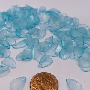 Könnte beinhalten: Ein Haufen durchsichtiger, hellblauer, tropfenförmiger Perlen mit einem Penny als Größenvergleich. Die Perlen haben eine leicht schimmernde Oberfläche.