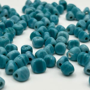 May include: A collection of turquoise-colored beads with various shapes, including rounded, square, and teardrop forms. Each bead has a hole for stringing. The beads are scattered on a white surface, creating a textured appearance.
