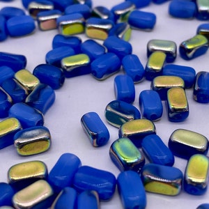 Può includere: Un insieme di perline blu rettangolari con bordi arrotondati. Alcune perline hanno una finitura metallica iridescente nei toni dell'oro e del verde. Le perline sono sparse su una superficie bianca, per la creazione di gioielli.