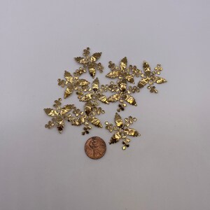 May include: A group of ten gold metal leaf-shaped embellishments, each with a delicate, intricate design. The embellishments are scattered on a white surface, with a penny coin for scale.