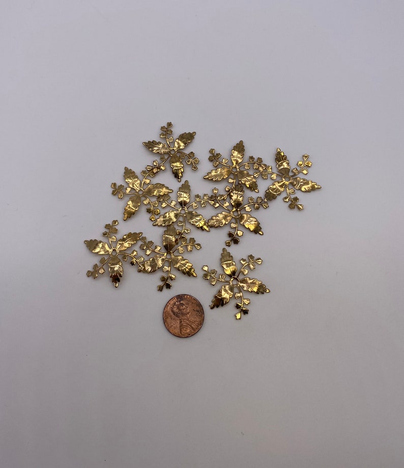 May include: A group of ten gold metal leaf-shaped embellishments, each with a delicate, intricate design. The embellishments are scattered on a white surface, with a penny coin for scale.