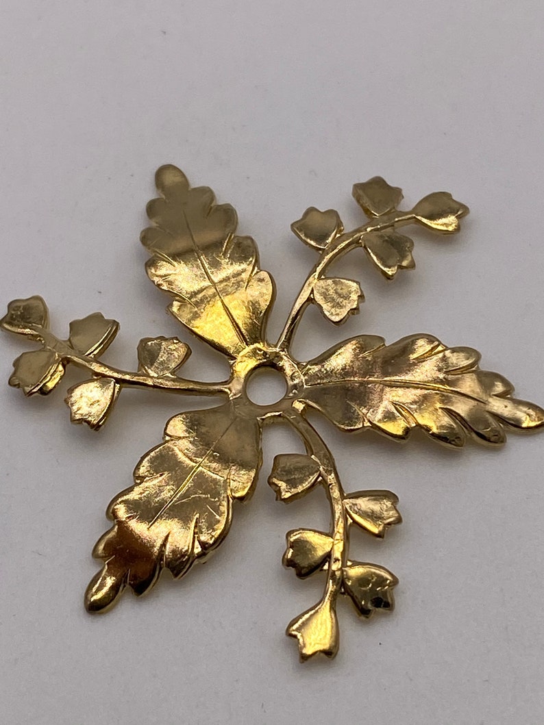 May include: Gold-toned metal pendant with a floral design. The pendant features a central hole and five branches with leaves and small flowers.