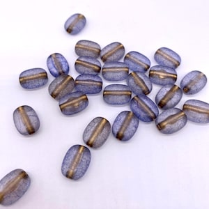 Può includere: Perline ovali blu traslucide con una striscia dorata al centro. Le perline hanno una texture screpolata e sono sparse su una superficie bianca. Queste perline sono adatte per la creazione di gioielli.