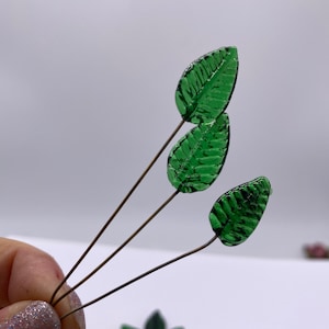 May include: A set of green glass leaves on wire stems. The leaves have a veined texture and are perfect for crafting.