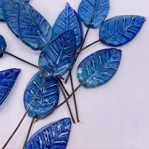 Könnte beinhalten: Eine Sammlung blauer Glasblatt-Ornamente mit detaillierten Adern. Jedes Blatt ist an einem dünnen, braunen Drahtstiel befestigt. Die Blätter sind ein leuchtendes, durchscheinendes Blau und sind vor einem weißen Hintergrund angeordnet.