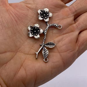 Puede incluir: Encanto de metal plateado con dos flores y una rama de hojas con dos anillos de salto.