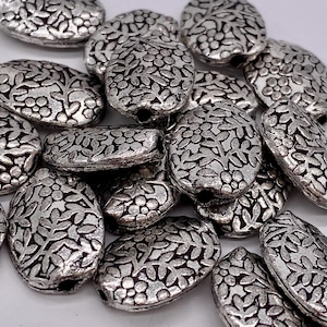 May include: A pile of silver oval-shaped beads with a floral design. The beads are approximately 2.5 cm long and have a hole in the centre for stringing.