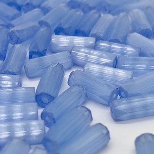 May include: A collection of light blue, cylindrical beads with a faceted, ribbed design. The beads are scattered across a white surface, creating a textured appearance. These beads are suitable for jewelry making and crafting projects.