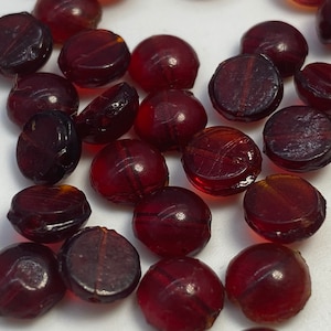 May include: A collection of dark red glass beads, each with a slightly different shape and texture. The beads are round and flat, with a smooth, polished surface.