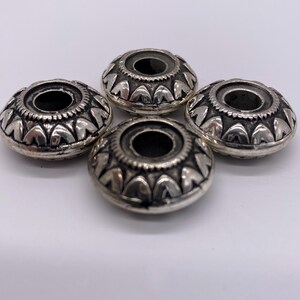 May include: Four silver-toned metal beads with a decorative design. Each bead has a circular hole in the center.