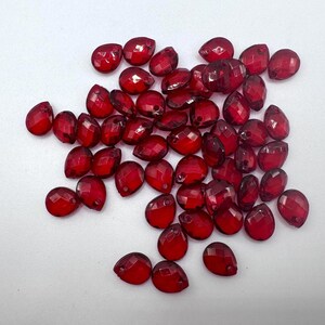 Può includere: Una collezione di piccole perline sfaccettate a forma di goccia di un colore rosso traslucido intenso. Ogni perlina presenta un foro praticato sulla punta superiore, progettato per l'infilatura. Le perline hanno una finitura superficiale lucida e riflettente.