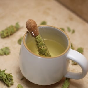 May include: A white mug with a light brown rim filled with a pale yellow liquid. A glass tube with a cork stopper containing green herbs sits in the mug. Green herbs are scattered around the mug on a light brown surface.