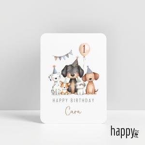 May include: A white birthday card featuring watercolor illustrations of five dogs wearing party hats. A banner and a balloon with the number 1 are above the dogs. The text "HAPPY BIRTHDAY Cara" is below the dogs.