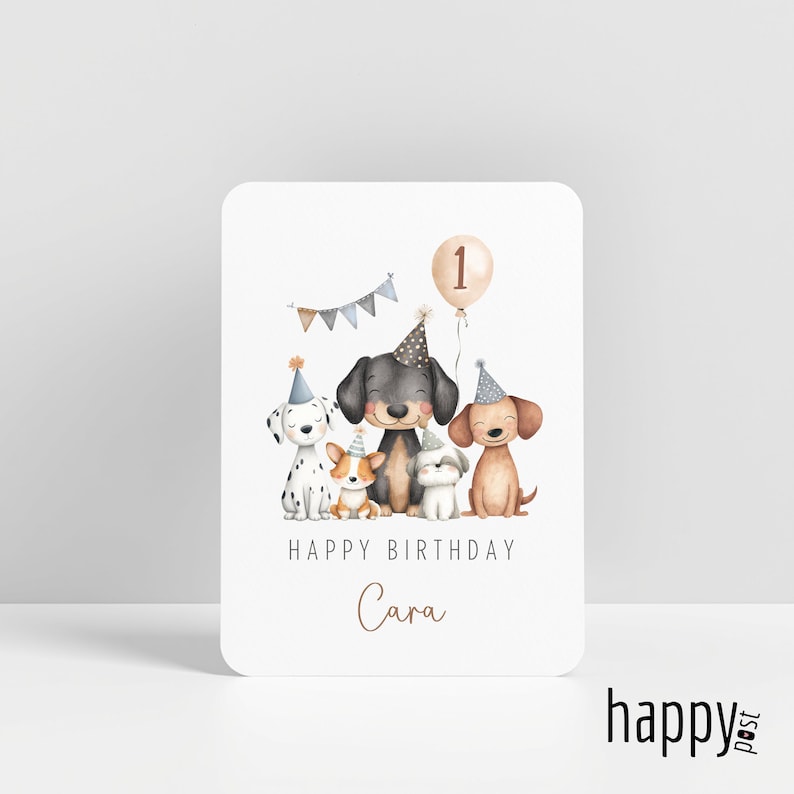 May include: A white birthday card featuring watercolor illustrations of five dogs wearing party hats. A banner and a balloon with the number 1 are above the dogs. The text "HAPPY BIRTHDAY Cara" is below the dogs.