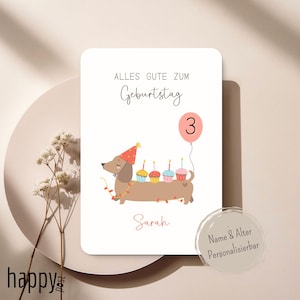 May include: A birthday card with a dachshund illustration. The card features the text "Alles Gute Zum Geburtstag" and "3" on a pink balloon. The dachshund wears a party hat and carries cupcakes with lit candles. The name "Sarah" is written below.
