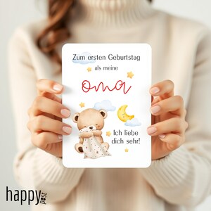 May include: A greeting card with the text "Zum ersten Geburtstag als meine Oma" and "Ich liebe dich sehr!" features a teddy bear, moon, and stars illustration. The card is held by a person.