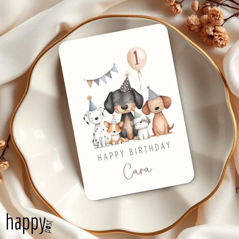 May include: A birthday card featuring a watercolor illustration of five dogs wearing party hats. A balloon with the number 1 floats above them. The words "HAPPY BIRTHDAY Cara" are written below the dogs. The card is on a white plate.