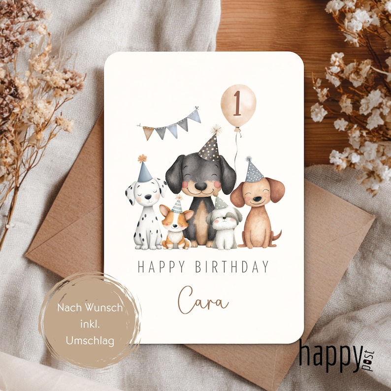 May include: A birthday card featuring a watercolor illustration of six cartoon dogs wearing party hats. The card reads "Happy Birthday Cara" with a balloon displaying the number "1". The card is on a neutral background with a brown envelope.