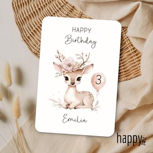 May include: A white birthday card with a watercolor illustration of a fawn wearing a floral crown and holding a balloon with the number 3. The card reads "Happy Birthday" and "Emilia". The bottom right corner has the word "happy post".