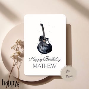 Può includere: Biglietto di auguri bianco con un'illustrazione ad acquerello di una chitarra acustica nera. Il biglietto recita "Happy Birthday MATHEW". Un piccolo adesivo circolare con la scritta "Mit Name" si trova in basso a destra.