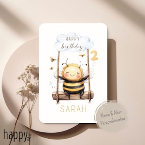 May include: A birthday card featuring a cartoon bee on a swing. The bee has a yellow and black striped body, with the number "2" and the name "SARAH" in gold. The card has the words "HAPPY BIRTHDAY" above the bee.