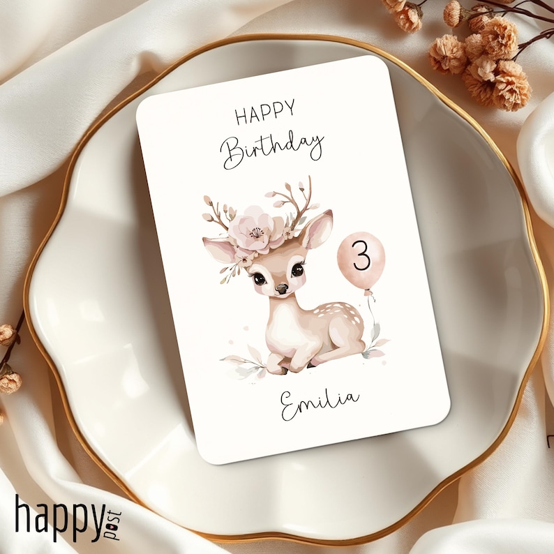 May include: A white birthday card with a watercolor illustration of a fawn wearing a floral crown and holding a pink balloon with the number 3. The card reads "Happy Birthday" and "Emilia". The card is on a white plate with a gold rim.