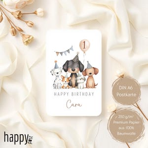 May include: A birthday card featuring a group of cartoon dogs wearing party hats. The card has the text "HAPPY BIRTHDAY Cara" and a balloon with the number 1. The card is white with a beige background and the words "DIN A6 Postkarte" and "350 g/m&sup2; Premium Papier aus 100% Baumwolle".
