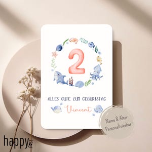 Op de afbeelding: Verjaardagskaart met een aquarelontwerp. Het toont een koraalrood nummer 2, omringd door zeedieren. De tekst "ALLES GUTE ZUM GEBURTSTAG" en een naam in rood schrift zijn inbegrepen. Een kleine, ronde sticker zegt "Name & Alter Personalisierbar".