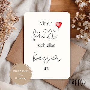 May include: A cream-coloured greeting card with the German text "Mit dir fühlt sich alles besser an" and a red heart balloon. The card is placed on a neutral surface with a brown envelope.