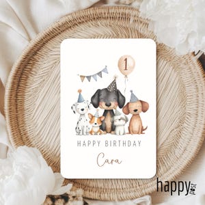 May include: A rectangular birthday card featuring a watercolor illustration of six cartoon dogs wearing party hats. The card reads "Happy Birthday Cara" with a balloon and banner. The card is on a woven surface.