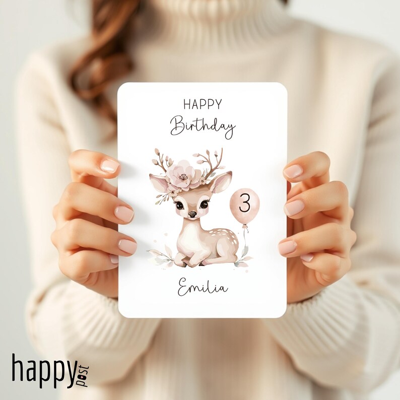 May include: A birthday card featuring a watercolor illustration of a fawn with floral antlers and a pink balloon with the number 3. The card reads "Happy Birthday" and "Emilia". The card is held by a person.