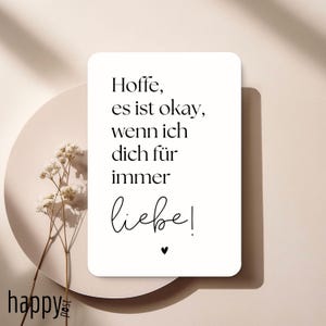 Peut inclure: Une carte rectangulaire blanche avec le texte "Hoffe, es ist okay, wenn ich dich für immer liebe!" en script noir. Un petit cœur noir est en dessous du texte. La carte est sur une assiette beige avec des fleurs séchées.