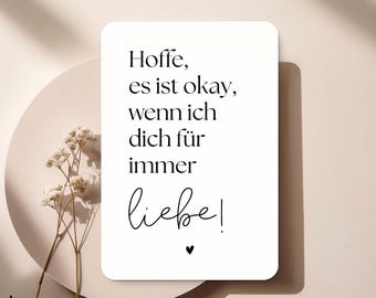 Op de afbeelding: Een witte rechthoekige kaart met de tekst "Hoffe, es ist okay, wenn ich dich für immer liebe!" in zwart script. Een klein zwart hart staat onder de tekst. De kaart ligt op een beige bord met gedroogde bloemen.